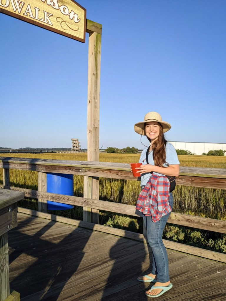girl with long brown hair wearing a sun hat smiling with the sun in her eyes standing on a boardwalk with marsh grass and clear blue sky in the background.