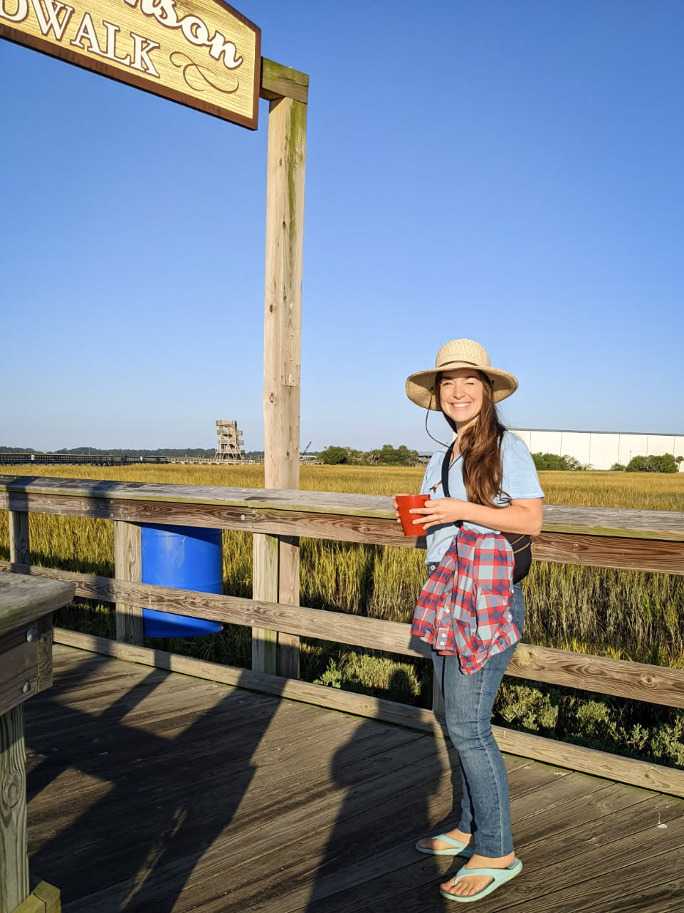 girl with long brown hair wearing a sun hat smiling with the sun in her eyes standing on a boardwalk with marsh grass and clear blue sky in the background.