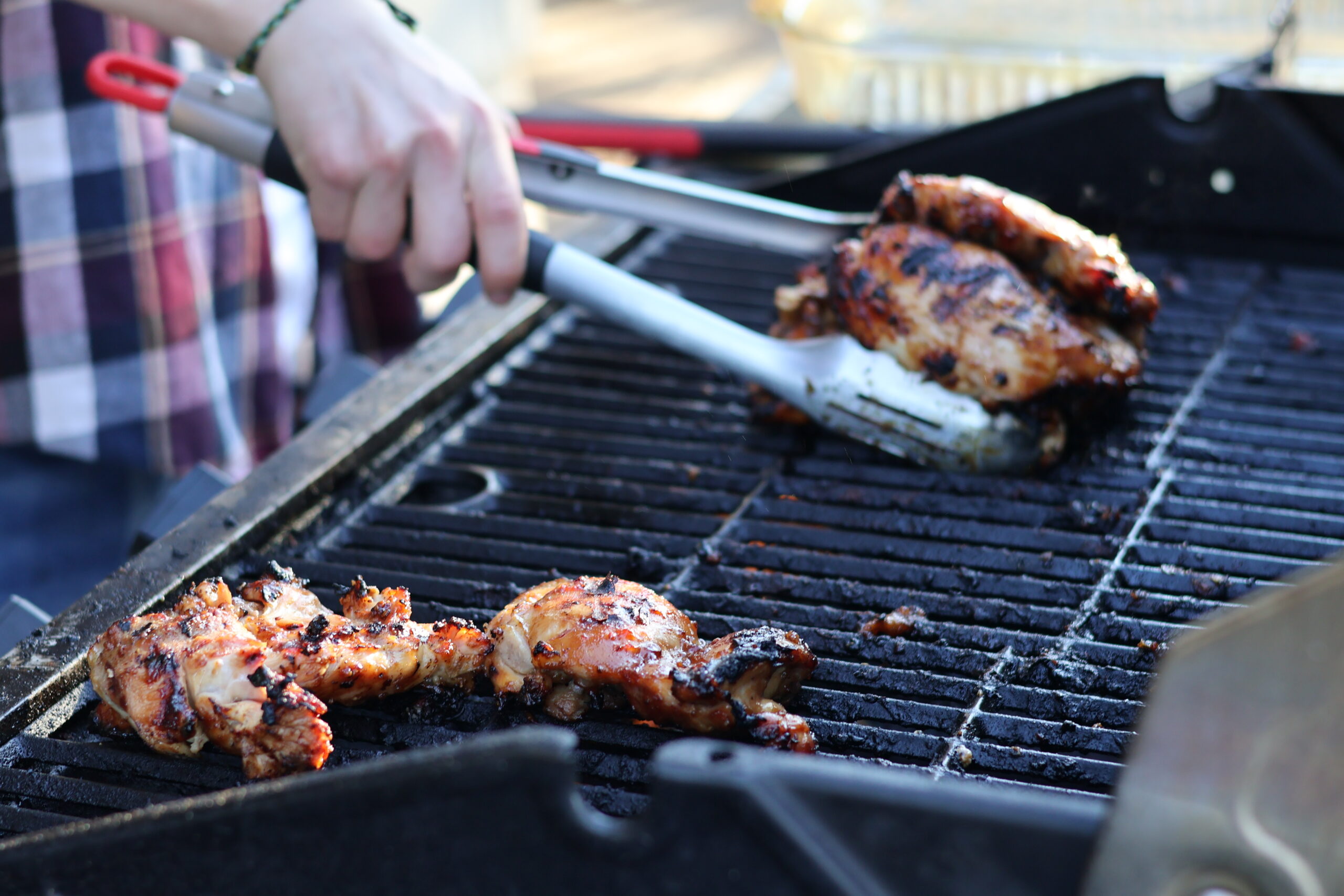 young man in plaid shirt grabbing grilled teriyaki chicken thighs off of a hot grill with tongs